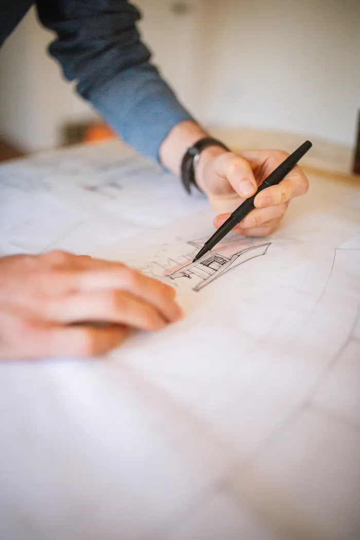 Overhead photograph of the studio desk: two hands inking a building elevation over blueprints