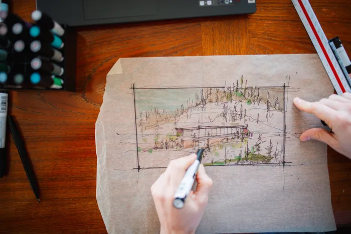 Overhead photograph of the studio desk: a hand sketching a building elevation in marker on kraft paper