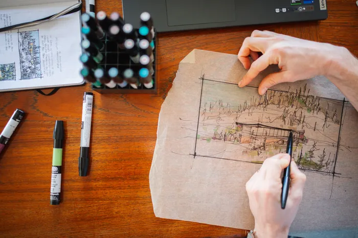 Overhead photograph of the studio desk: two hands drawing a building perspective in marker on kraft paper