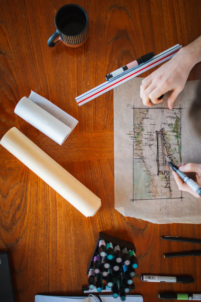 Overhead photograph of the studio desk: a hand sketching over coffee, rolled paper, and a marker set